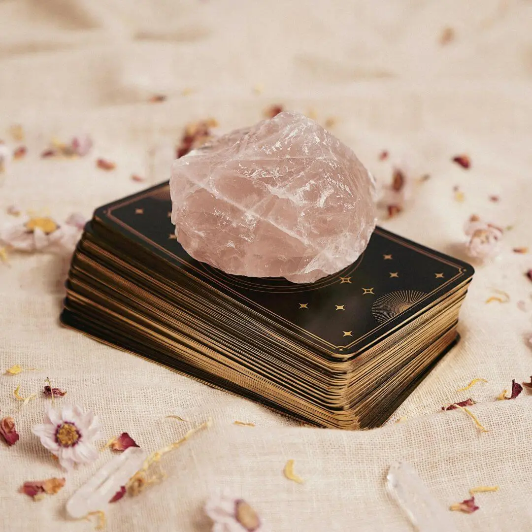 Quartz rose posé sur un jeu de cartes divinatoires dorées, au milieu de pétales et fleurs séchées dans une atmosphère douce et spirituelle.