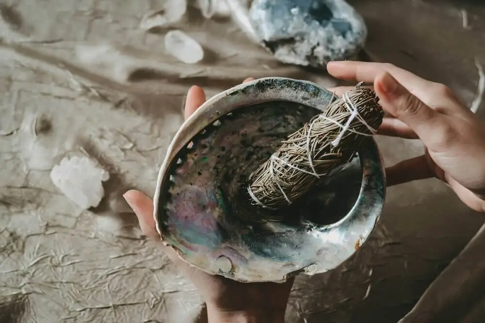 nettoyer-purifier-oracle-rituel-sauge Mains tenant une coquille nacrée contenant un bâton de sauge pour nettoyer et purifier un oracle divinatoire lors d’un rituel spirituel.